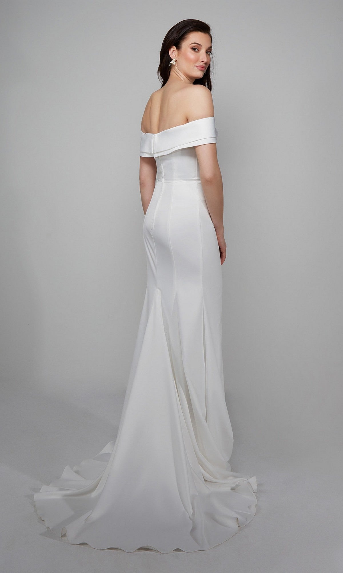 A woman stands against a plain gray background, modeling the Long White Dress: Alyce 7058—an elegant off-the-shoulder gown with a fitted bodice, floor-length skirt, and subtle train.